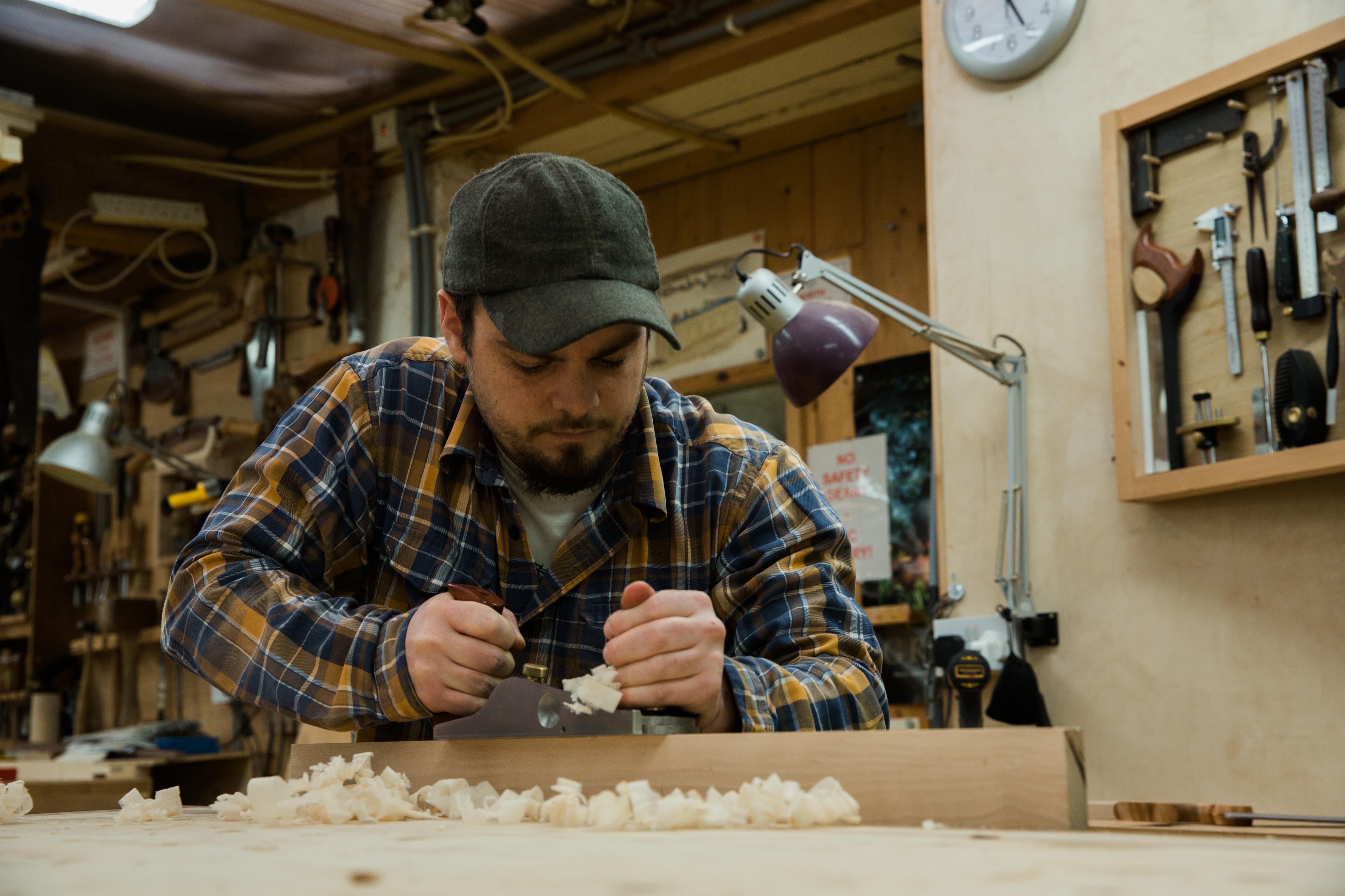 John Grillo sitting at his workbench, using a hand plane to even out a surface, epitomizing craftsmanship and dedication